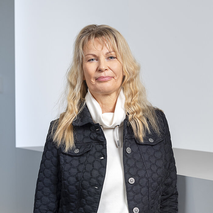 Porträtaufnahme einer Frau mit langem, blondem, gewelltem Haar und blauen Augen. Sie trägt eine schwarze, gesteppte Jacke über einem weißen Rollkragenpullover und blickt direkt in die Kamera. Der Hintergrund ist eine schlichte, helle Wand.