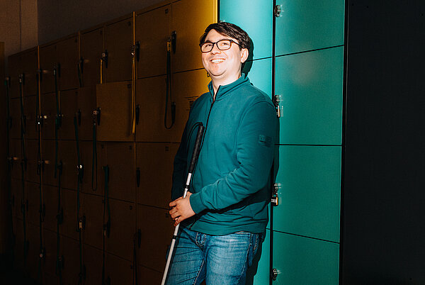 Porträtaufnahme eines lächelnden jungen Mannes mit Brille, der an einer Wand mit Spinden lehnt. Er trägt einen blaugrünen Troyer und Jeans. In seiner rechten Hand hält er einen zusammengeklappten Blindenstock. Die Spinde im Hintergrund sind teils gelb, teils in einem kräftigen Türkis gehalten.