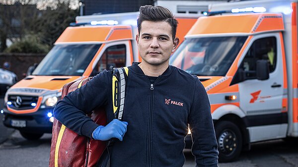 Porträtaufnahme eines jungen Sanitäters oder Rettungsassistenten, der vor zwei Krankenwagen steht. Er trägt eine dunkle Einsatzjacke mit einem roten "FALCK" Logo, blaue Einweghandschuhe und eine rote/gelbe Notfalltasche über der Schulter. Er blickt direkt in die Kamera mit ernstem Ausdruck. Die Krankenwagen im Hintergrund sind weiß mit orangefarbenen Streifen und eingeschaltetem Blaulicht.