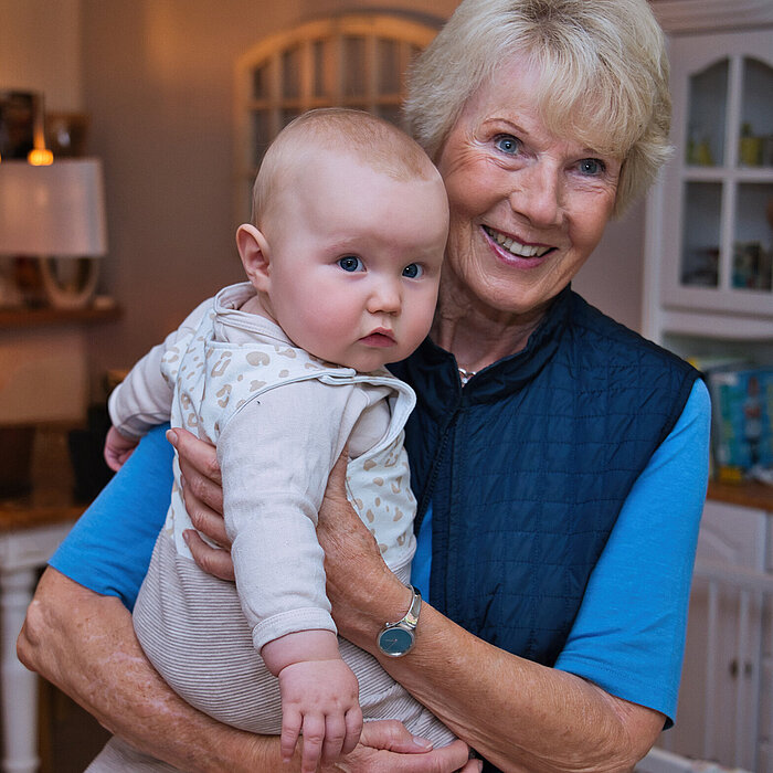 Porträtaufnahme einer lächelnden, älteren Frau mit hellem Haar, die ein kleines Baby im Arm hält. Die Frau trägt eine dunkelblaue, gesteppte Weste über einem blauen Shirt. Das Baby trägt einen hellen Strampler und schaut nach vorne. Im Hintergrund ist ein Innenraum, wahrscheinlich eine Küche oder ein Wohnzimmer, mit unscharfen Schränken und einer Lampe zu sehen.