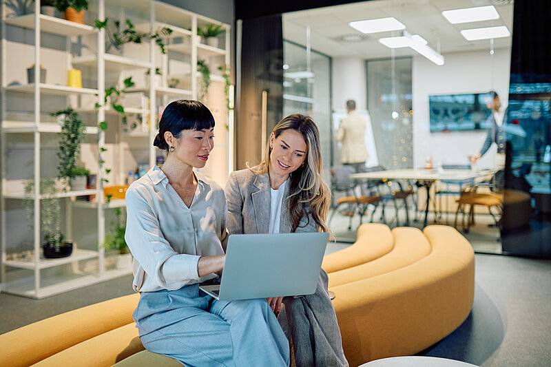 Zwei Frauen sitzen nebeneinander auf einem geschwungenen, ockerfarbenen Polstermöbel in einer modernen Bürolandschaft und blicken gemeinsam auf einen Laptop. Die linke Frau mit dunklen, hochgesteckten Haaren deutet auf den Bildschirm, während die rechte Frau mit langen blonden Haaren aufmerksam zusieht. Im Hintergrund ist ein verglaster Konferenzraum mit weiteren Personen sowie ein weißes Regalsystem mit vielen grünen Zimmerpflanzen zu sehen.