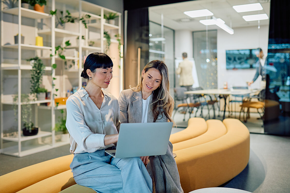Zwei Frauen sitzen nebeneinander auf einem geschwungenen, ockerfarbenen Polstermöbel in einer modernen Bürolandschaft und blicken gemeinsam auf einen Laptop. Die linke Frau mit dunklen, hochgesteckten Haaren deutet auf den Bildschirm, während die rechte Frau mit langen blonden Haaren aufmerksam zusieht. Im Hintergrund ist ein verglaster Konferenzraum mit weiteren Personen sowie ein weißes Regalsystem mit vielen grünen Zimmerpflanzen zu sehen.