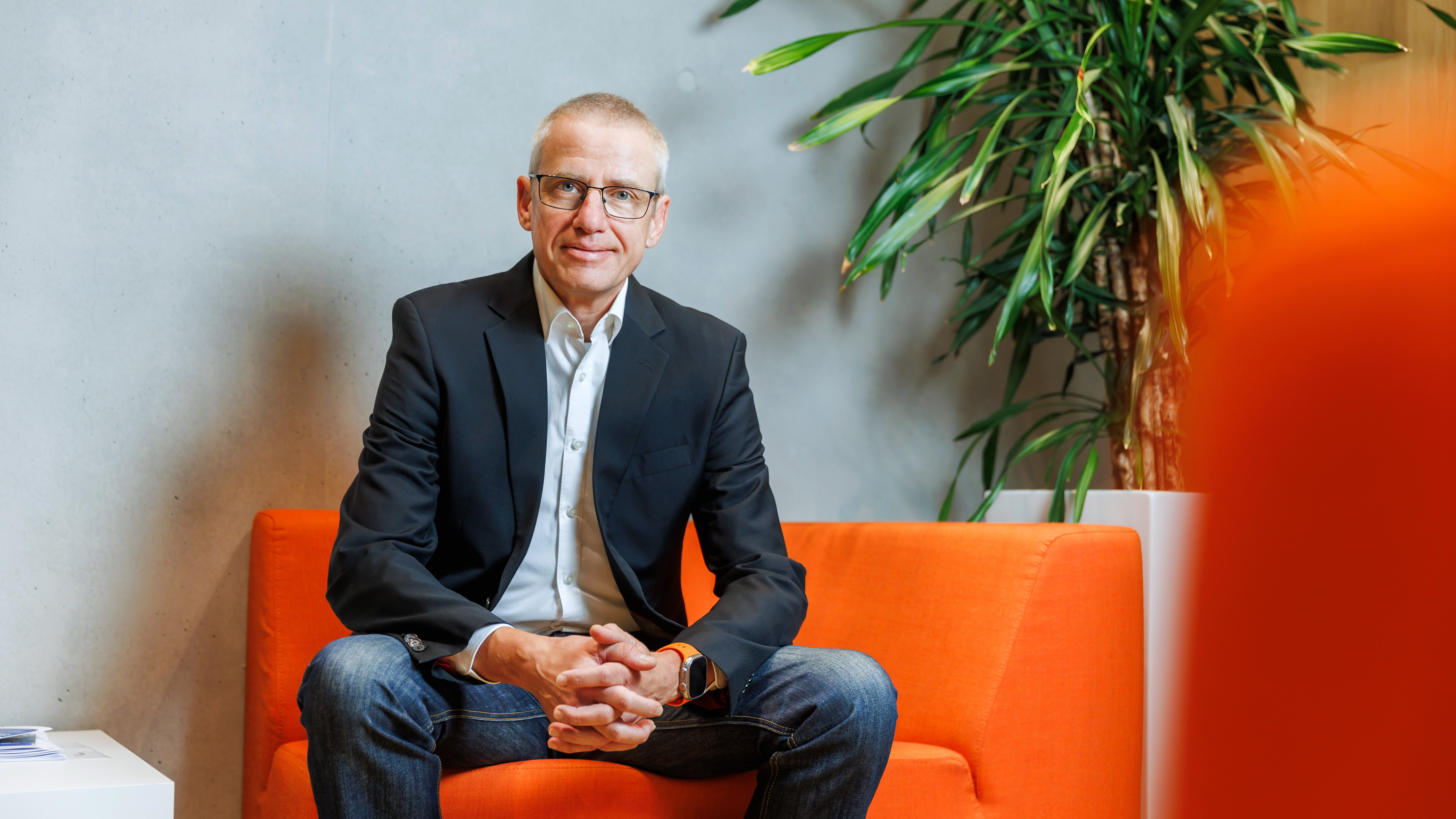 Ein Mann in einem schwarzen Sakko und Jeans sitzt auf einem orangefarbenen Sofa. Er hat die Hände gefaltet und lächelt leicht. Im Hintergrund steht eine große Pflanze.