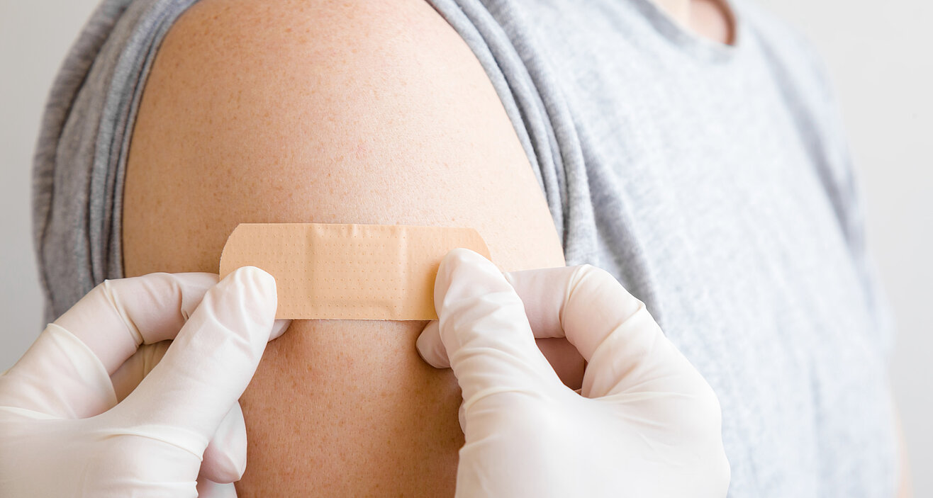 Foto: Ein Plaster wird auf einen entblößten Oberarm geklebt. Die Person, die das Pflaster aufklebt, trägt weiße Laborhandschuhe.