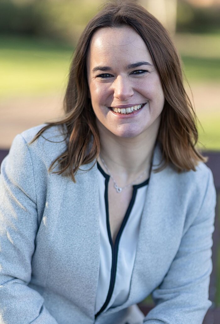 Porträtaufnahme einer lächelnden Frau mit schulterlangem braunem Haar. Sie trägt einen hellgrauen Blazer mit dunkler Verzierung am Kragen und eine feine Halskette. Sie blickt direkt in die Kamera. Der Hintergrund ist unscharf und zeigt eine grüne Außenaufnahme, wahrscheinlich in einem Park oder Garten.