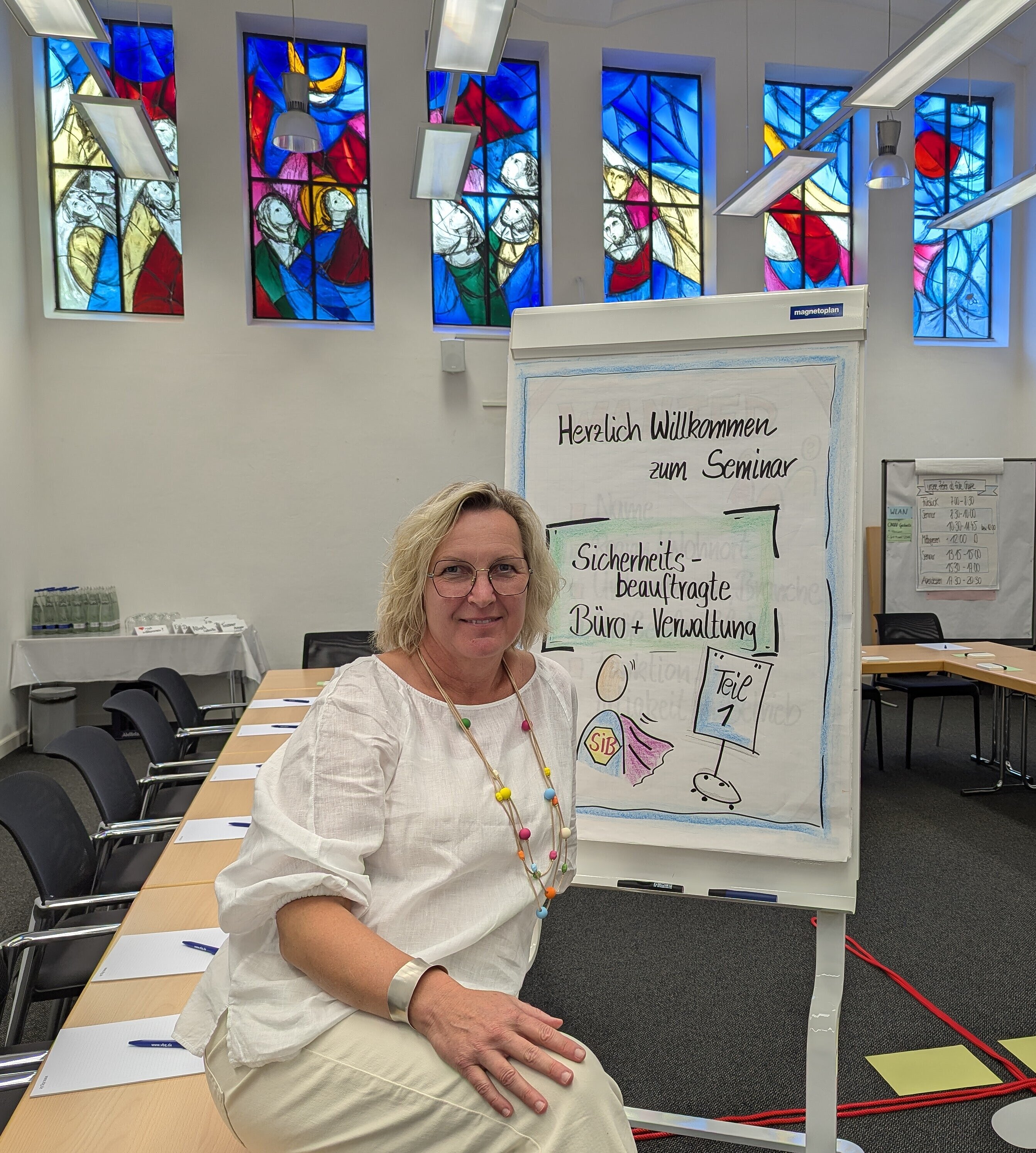 Eine Frau sitzt lächelnd neben einem Flipchart in einem Seminarraum. Auf dem Flipchart steht "Herzlich Willkommen zum Seminar Sicherheitsbeauftragte Büro + Verwaltung Teil 1". Der Raum ist mit bunten, kunstvollen Glasfenstern dekoriert. Im Hintergrund sind Tische mit Stühlen, Schreibblöcken und Stiften vorbereitet.