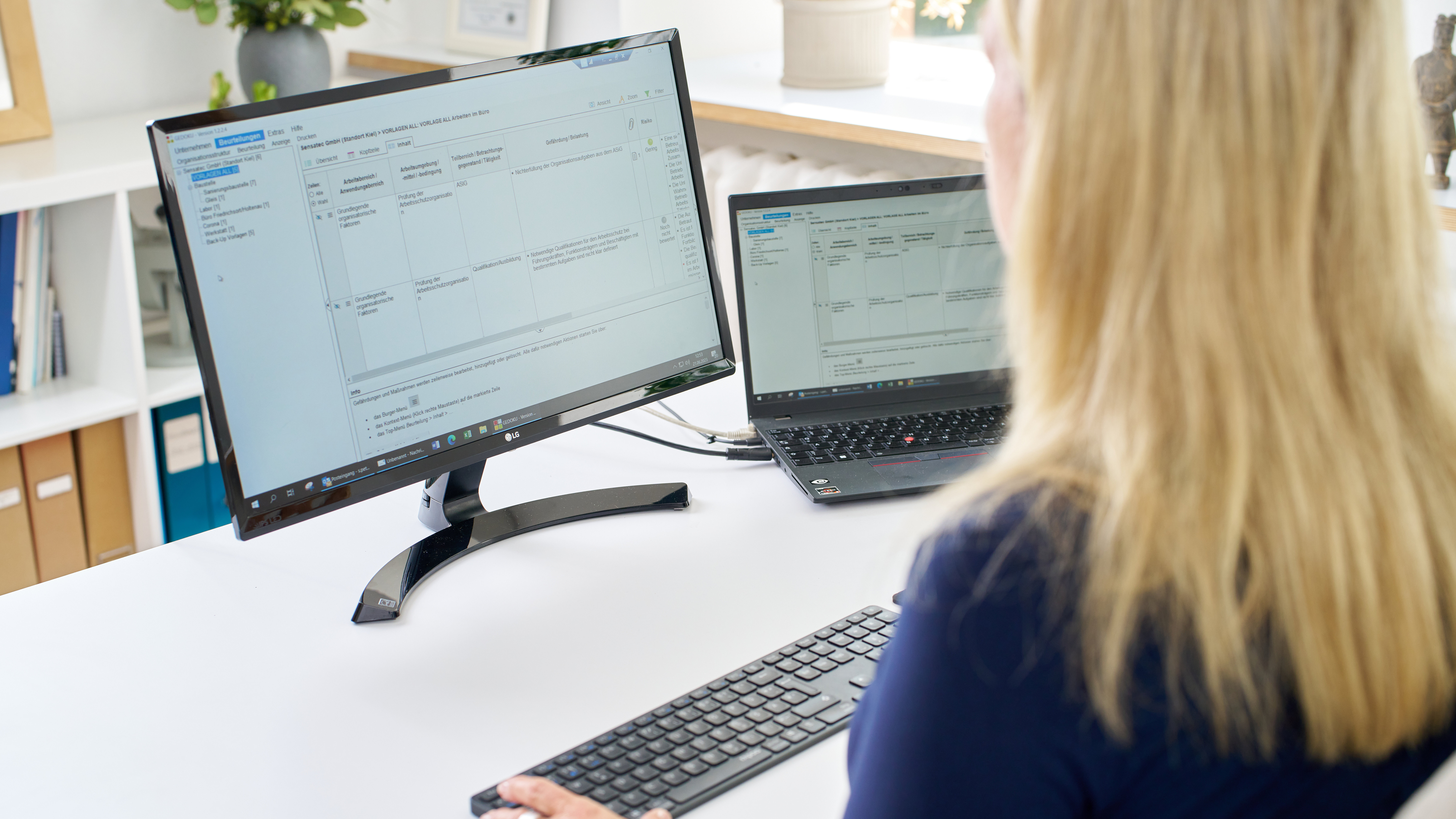 Über die Schulter einer blinden Frau schauend, sieht man auf einem weißen Schreibtisch einen Bildschirm mit einer geöffneten Software. Rechts daneben steht auch ein schwarzer Laptop. Auch hier ist eine Software geöffnet. 