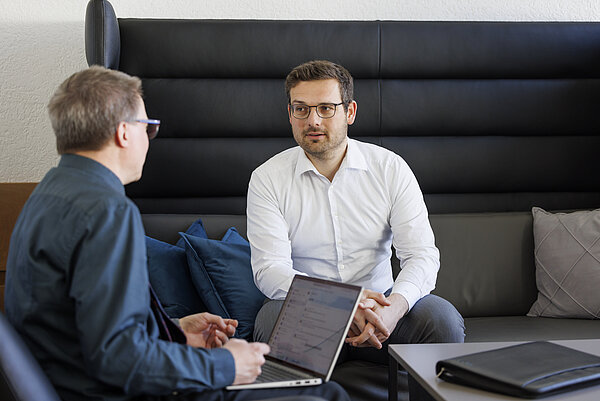 Zwei Männer sitzen in einem modernen Besprechungsbereich und unterhalten sich. Der Mann rechts trägt ein weißes Hemd, eine Brille und sitzt auf einem dunkelgrauen Sofa mit hoher Rückenlehne. Der Mann links sitzt ihm schräg gegenüber, trägt ein dunkles Hemd und eine Brille und hat einen geöffneten Laptop auf dem Schoß. Zwischen ihnen liegen blaue Kissen. Die beiden Männer blicken einander an.