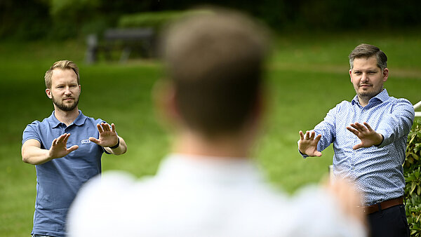 Zwei Männer stehen im Freien auf einer grünen Wiese und führen synchrone Übungen aus, möglicherweise Qigong oder eine Entspannungsübung. Sie strecken beide Arme nach vorne, die Handflächen nach außen. Der Mann links trägt ein blaues Poloshirt. Der Mann rechts trägt ein hellblaues Hemd. Ein dritter Mann steht mit dem Rücken zur Kamera im unscharfen Vordergrund und führt ebenfalls die Übung aus. Der Hintergrund ist ein grüner Park oder eine Wiese.