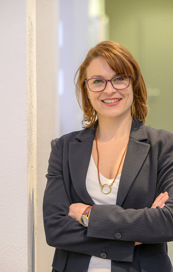 Porträtaufnahme einer lächelnden Frau mit schulterlangem rotem oder braunem Haar und Pony. Sie trägt eine Brille mit dunklem Rahmen, einen dunklen Blazer über einem weißen Oberteil und eine lange Kette mit einem großen runden Anhänger. Sie hat die Arme verschränkt und lehnt sich an eine helle Wand. Der Hintergrund ist ein schlichter Innenraum.