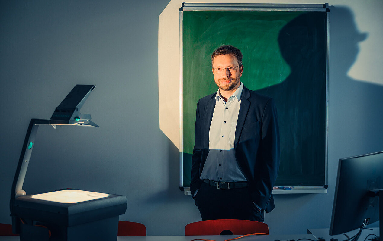 Ein Mann in einem Anzug steht in einem Raum mit einem Overheadprojektor und einer grünen Tafel. Die Beleuchtung erzeugt einen Schatten an der Wand.