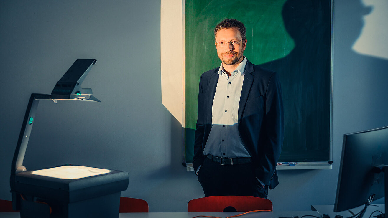 Ein Mann in einem Anzug steht in einem Raum mit einem Overheadprojektor und einer grünen Tafel. Die Beleuchtung erzeugt einen Schatten an der Wand.