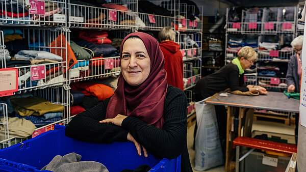 Eine lächelnde Frau mittleren Alters mit einem weinroten Hidschāb sitzt oder lehnt sich auf einen blauen Tisch oder Tresen und schaut in die Kamera. Sie trägt ein dunkles Oberteil. Im Hintergrund ist ein Lagerraum oder eine Kleiderkammer zu sehen, mit weißen Drahtregalen, die mit gestapelten Kleidungsstücken gefüllt sind. Andere Personen arbeiten im unscharfen Hintergrund.