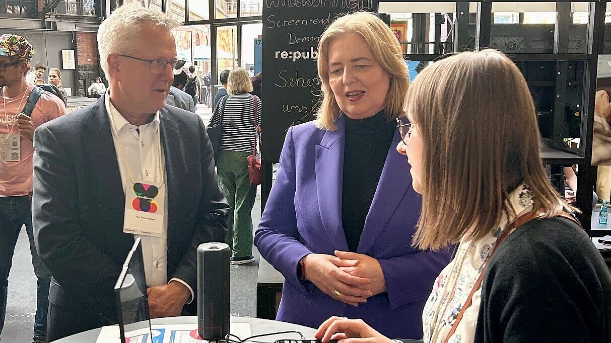 Drei Personen stehen bei einer Veranstaltung in einem Gespräch zusammen. Links steht ein älterer Mann mit grauem Haar und Brille in einem Anzug. In der Mitte steht eine blonde Frau in einem lila Blazer über einem schwarzen Rollkragenpullover, die mit den anderen beiden Personen spricht. Rechts ist eine Frau mit braunem Haar und Brille, die teilweise angeschnitten ist und zu der Frau in Lila spricht. Im Hintergrund sind weitere Personen und eine Tafel mit dem Wort "re:publica" und anderen Texten zu sehen, was auf eine Konferenz hindeutet.