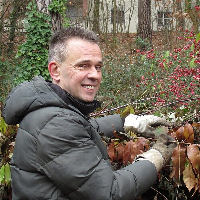 Ein lächelnder Mann mittleren Alters mit kurzen, dunklen Haaren arbeitet draußen in einem Garten oder Park. Er trägt eine dicke, dunkle Winterjacke und Arbeitshandschuhe. Er schneidet oder bindet etwas an einer Hecke oder einem Busch mit roten Beeren und braunen Blättern. Im unscharfen Hintergrund sind weitere Bäume und möglicherweise ein Gebäude zu sehen.