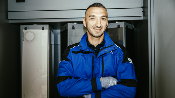  Porträtaufnahme eines lächelnden Mannes mittleren Alters, der eine blaue Arbeitsjacke mit schwarzen Akzenten trägt. Er hat die Arme verschränkt und trägt Arbeitshandschuhe. Er steht in einem technischen Raum oder vor Schaltschränken. Im Hintergrund sind silberne und dunkle, rechteckige Geräte oder Server-Racks zu sehen.