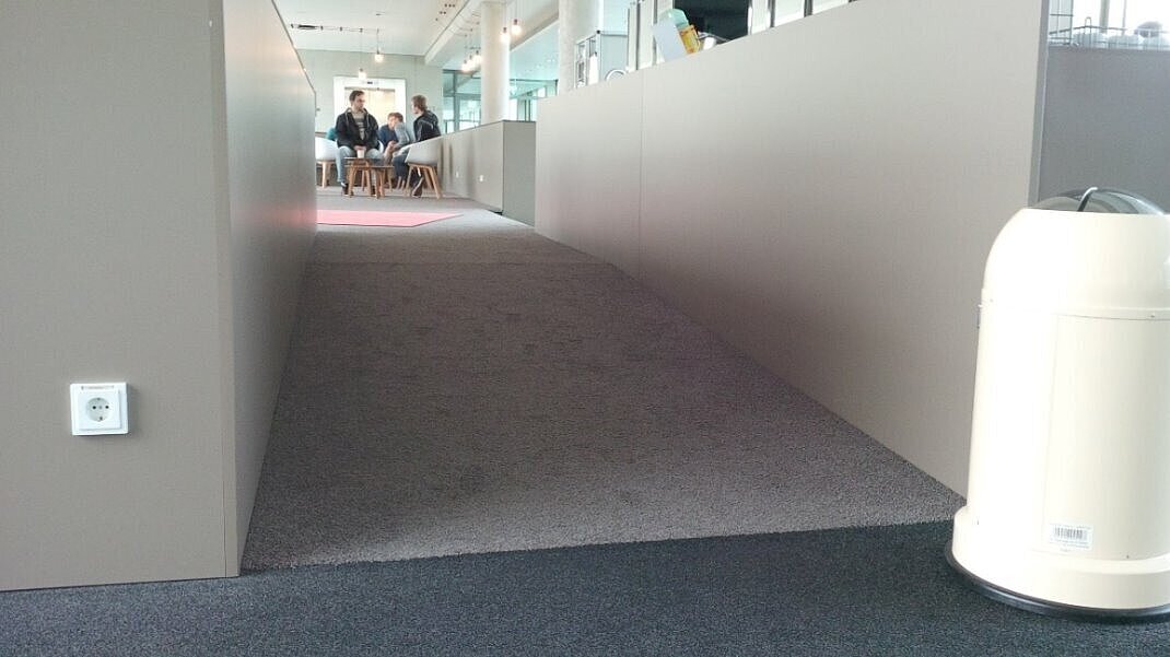 Eine flache, teppichbelegte Rampe führt in einem modernen Gebäude barrierefrei zu einem höher gelegenen Sitzbereich.
