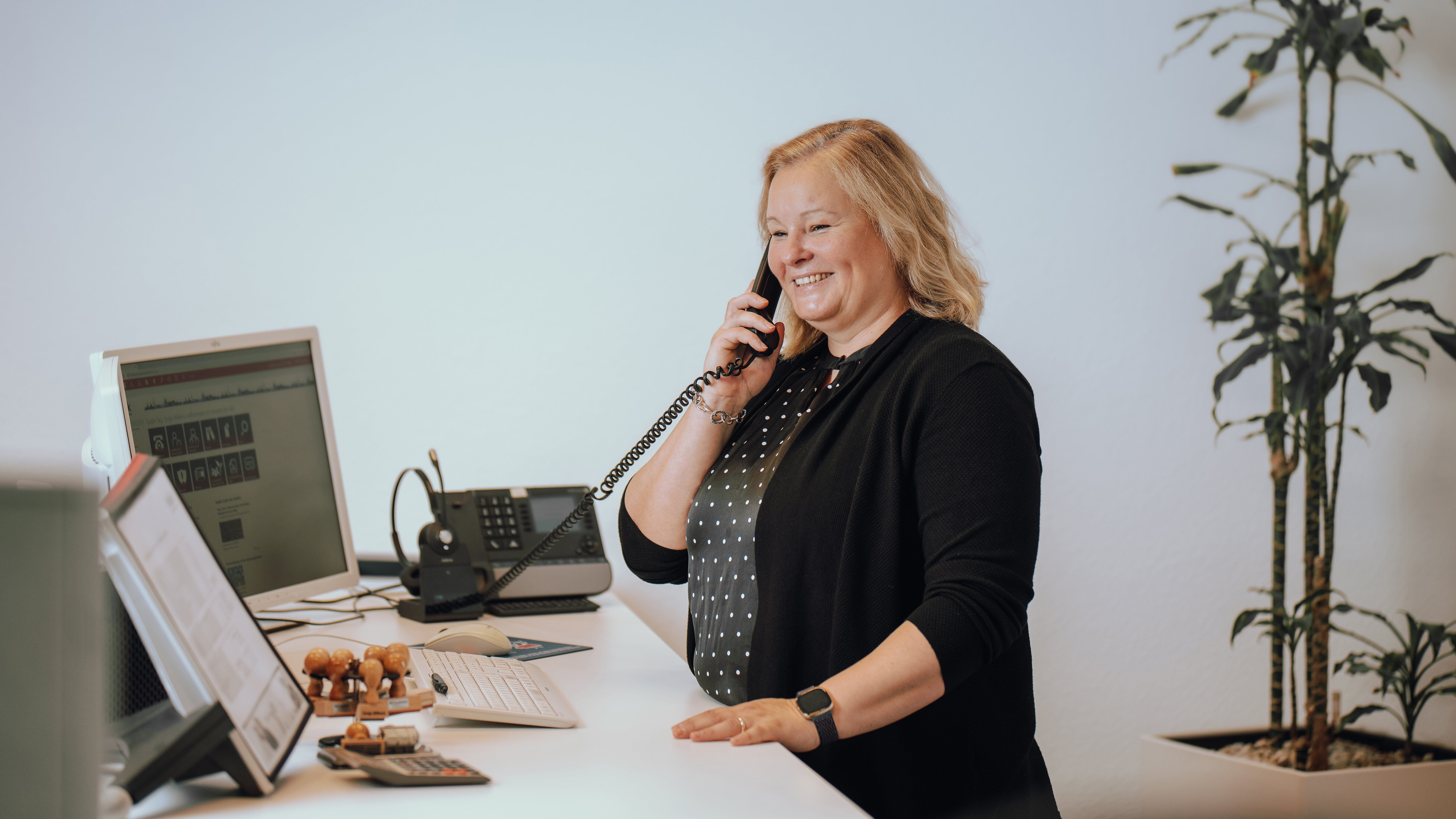 Eine Frau steht lächelnd an einem Schreibtisch und telefoniert. Vor ihr befinden sich ein Computerbildschirm, ein Telefon und einige Stempel. Im Hintergrund ist eine Pflanze zu sehen.