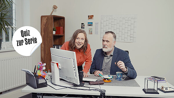 Zwei Personen sitzen oder stehen vor einem Computerbildschirm an einem Schreibtisch in einem Büro. Eine Frau mit langen braunen Haaren und einem orangefarbenen Oberteil beugt sich von links über den Bildschirm und lächelt in die Kamera. Rechts sitzt ein bärtiger Mann mit grauem Haar, der ein Sakko und ein helles Hemd trägt. Er blickt nachdenklich oder kritisch auf den Bildschirm und hält etwas Kleines in der Hand. Links im Bild befindet sich ein weißes Etikett mit dem schwarzen Text "Quiz zur Serie". Im Hintergrund ist ein Holzregal, ein Kalender und Fotos an der Wand zu sehen.