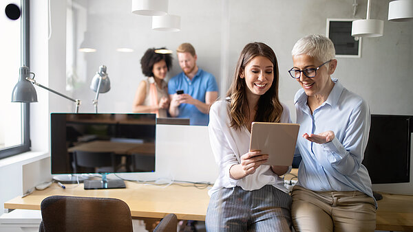 Zwei Frauen unterschiedlichen Alters sitzen nebeneinander in einem modernen Büro und schauen gemeinsam auf ein Tablet. Die jüngere Frau links hat lange, dunkle Haare und trägt eine weiße Bluse. Die ältere Frau rechts hat kurzes, helles Haar, eine Brille und trägt eine hellblaue Bluse, während sie mit einer Hand gestikuliert. Beide lächeln. Im unscharfen Hintergrund sind zwei weitere Kollegen bei der Arbeit oder im Gespräch zu sehen. Die Büroumgebung ist hell und modern mit Schreibtischen und Computern.