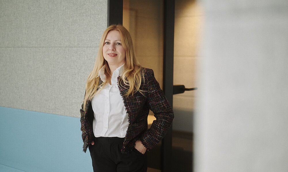 Porträt einer Frau mit langen blonden Haaren, die eine dunkle, bunt strukturierte Tweed-Jacke über einer weißen Bluse und eine dunkle Hose trägt. Sie steht lächelnd in einem modernen Bürogebäude mit den Händen in den Taschen.