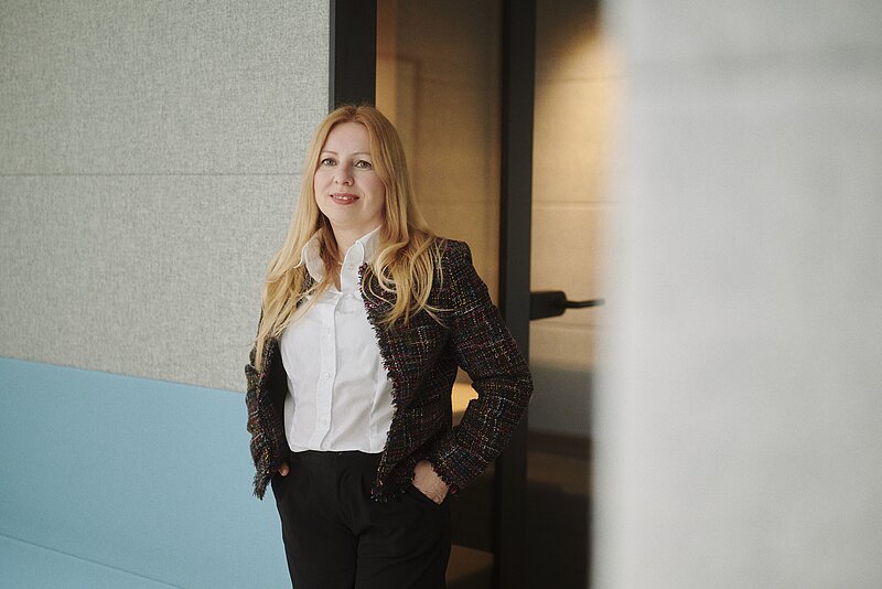 Porträt einer Frau mit langen blonden Haaren, die eine dunkle, bunt strukturierte Tweed-Jacke über einer weißen Bluse und eine dunkle Hose trägt. Sie steht lächelnd in einem modernen Bürogebäude mit den Händen in den Taschen.