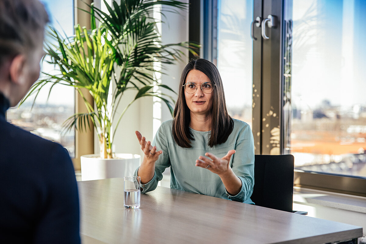 Eine Frau mit Brille und türkisfarbenem Oberteil sitzt an einem Tisch und gestikuliert beim Sprechen. Im Hintergrund sind große Fenster und eine Pflanze zu sehen. Eine weitere Person ist von hinten zu sehen.