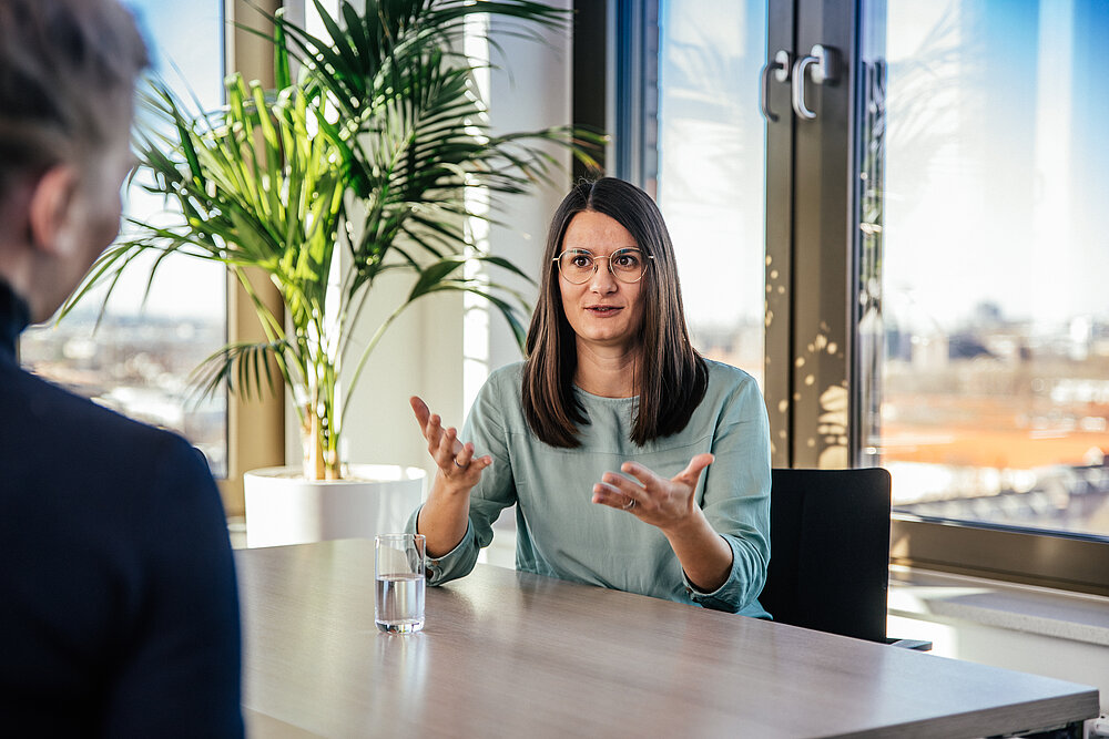 Eine Frau mit Brille und türkisfarbenem Oberteil sitzt an einem Tisch und gestikuliert beim Sprechen. Im Hintergrund sind große Fenster und eine Pflanze zu sehen. Eine weitere Person ist von hinten zu sehen.