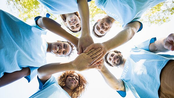 Eine Gruppe von sechs lächelnden, vielfältigen Personen steht im Freien in einem Kreis und streckt ihre Hände in die Mitte, wobei sie ihre Hände übereinander stapeln, als Symbol für Teamwork und Einheit. Alle tragen hellblaue oder türkise T-Shirts. Die Aufnahme ist von unten gemacht, wodurch die Gesichter der Personen in die Kamera schauen und von oben von hellem Licht beleuchtet werden. Im Hintergrund sind unscharf Bäume und der Himmel zu sehen.