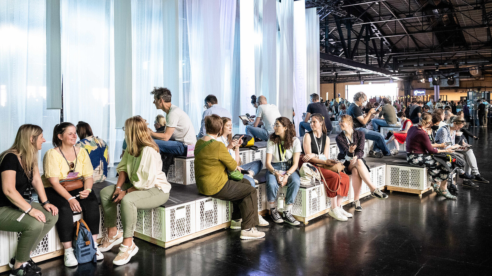 Foto: Gäste der re:publica sitzen in einer Messehalle auf Sitzmöbeln aus weißen Kisten und unterhalten sich.