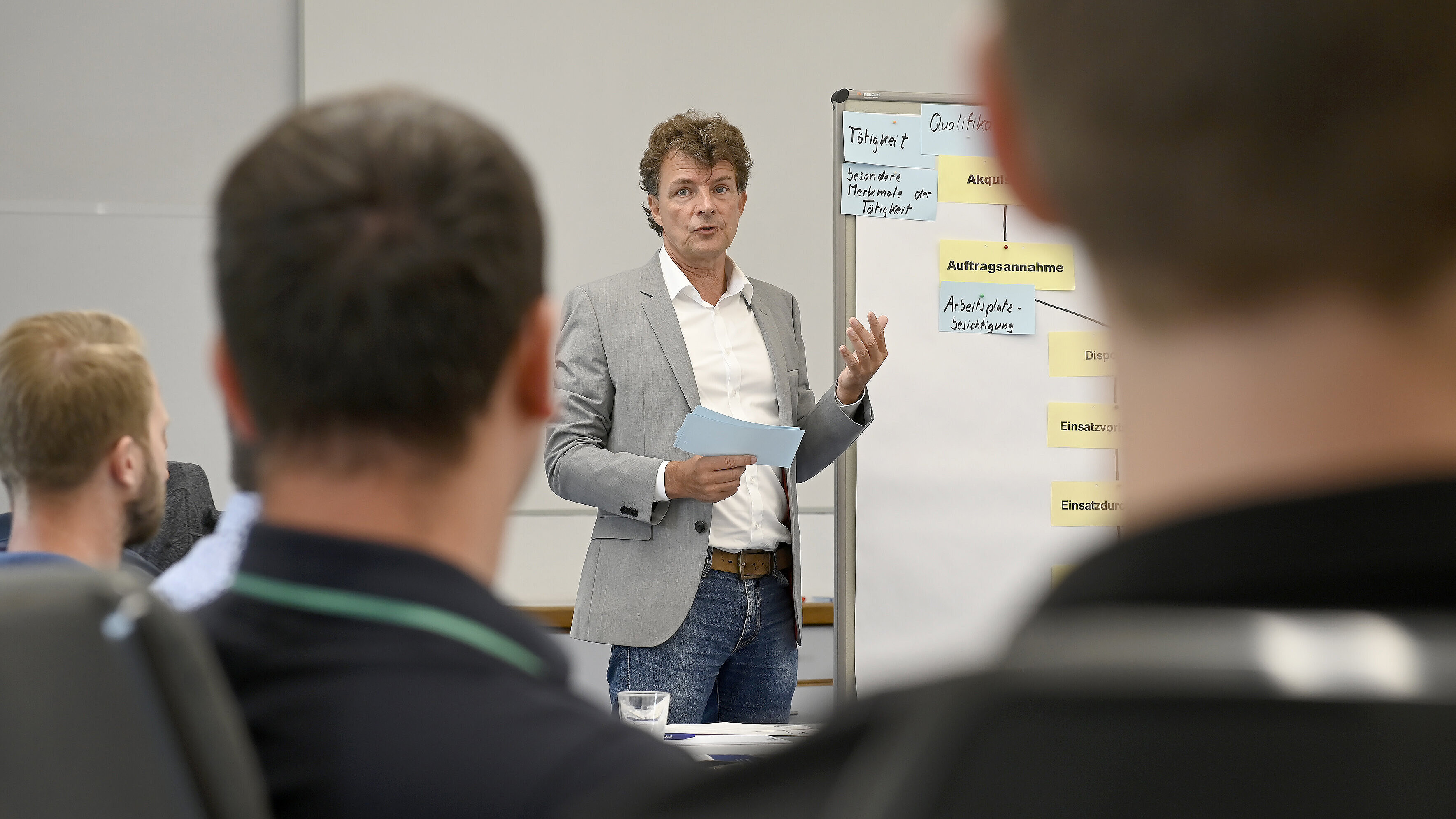 Rüdiger Hitzemann steht an einem Whiteboard und leitet das Seminar. Er hat braune Harre und trägt ein graues Sakko und ein weißes Hemd. Zwei Teilnehmende des Seminars sind von hinten zu sehen.