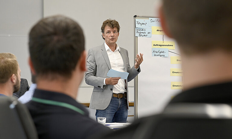 Rüdiger Hitzemann steht an einem Whiteboard und leitet das Seminar. Er hat braune Harre und trägt ein graues Sakko und ein weißes Hemd. Zwei Teilnehmende des Seminars sind von hinten zu sehen.