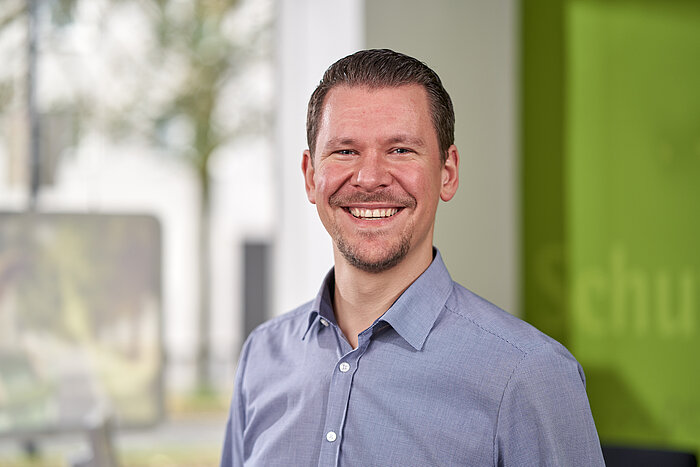 Porträtfoto von Alexander Boldt in seiner Versicherungsagentur. Er hat braunes Haar, einen leichten Bart und trägt ein hellblaues Hemd.