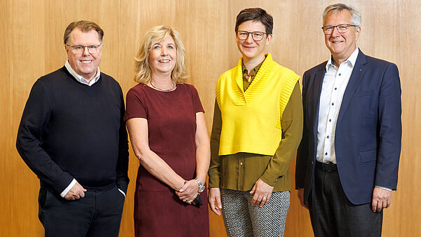 Eine Gruppe von vier Personen steht nebeneinander vor einer holzgetäfelten Wand und blickt in die Kamera. Von links nach rechts: 1. Ein Mann mit Brille in einem dunklen Pullover über einem weißen Hemd. 2. Eine lächelnde Frau mit blondem Haar in einem bordeauxroten Kleid. 3. Eine Frau mit kurzen dunklen Haaren und Brille, die eine gelbe Weste über einem dunklen Oberteil trägt. 4. Ein Mann mit Brille und hellem Haar in einem dunklen Sakko über einem hellen Hemd.
