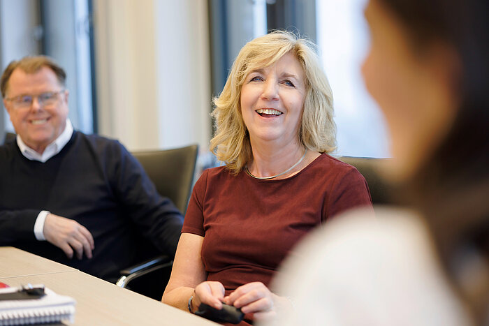 Eine blonde Frau sitzt in einem Besprechungsraum an einem Tisch, lacht und schaut nach rechts zu einer unscharfen Person im Vordergrund. Sie trägt ein bordeauxrotes Oberteil. Links sitzt ein Mann mit Brille und dunklem Pullover über einem weißen Hemd, der ebenfalls leicht lächelt. Die Szene ist hell, mit Fenstern im Hintergrund.