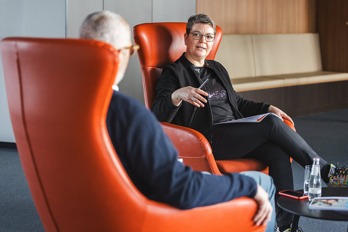 Zwei Personen sitzen sich in großen, orangefarbenen Ohrensesseln in einer modernen Büroumgebung gegenüber und führen ein Gespräch. Die Frau im Hintergrund trägt eine Brille und eine schwarze Jacke, hält einen Stift in der Hand und spricht gestikulierend. Der Mann im Vordergrund ist nur von hinten zu sehen. Auf einem kleinen Tisch neben ihnen stehen Wasserflaschen und ein Smartphone.