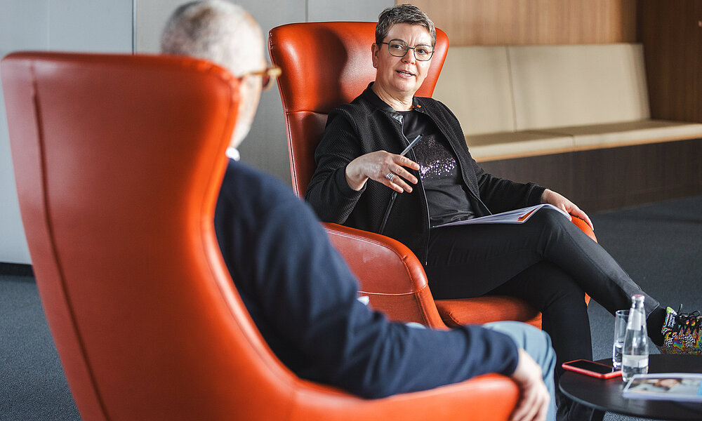 Zwei Personen sitzen sich in großen, orangefarbenen Ohrensesseln in einer modernen Büroumgebung gegenüber und führen ein Gespräch. Die Frau im Hintergrund trägt eine Brille und eine schwarze Jacke, hält einen Stift in der Hand und spricht gestikulierend. Der Mann im Vordergrund ist nur von hinten zu sehen. Auf einem kleinen Tisch neben ihnen stehen Wasserflaschen und ein Smartphone.