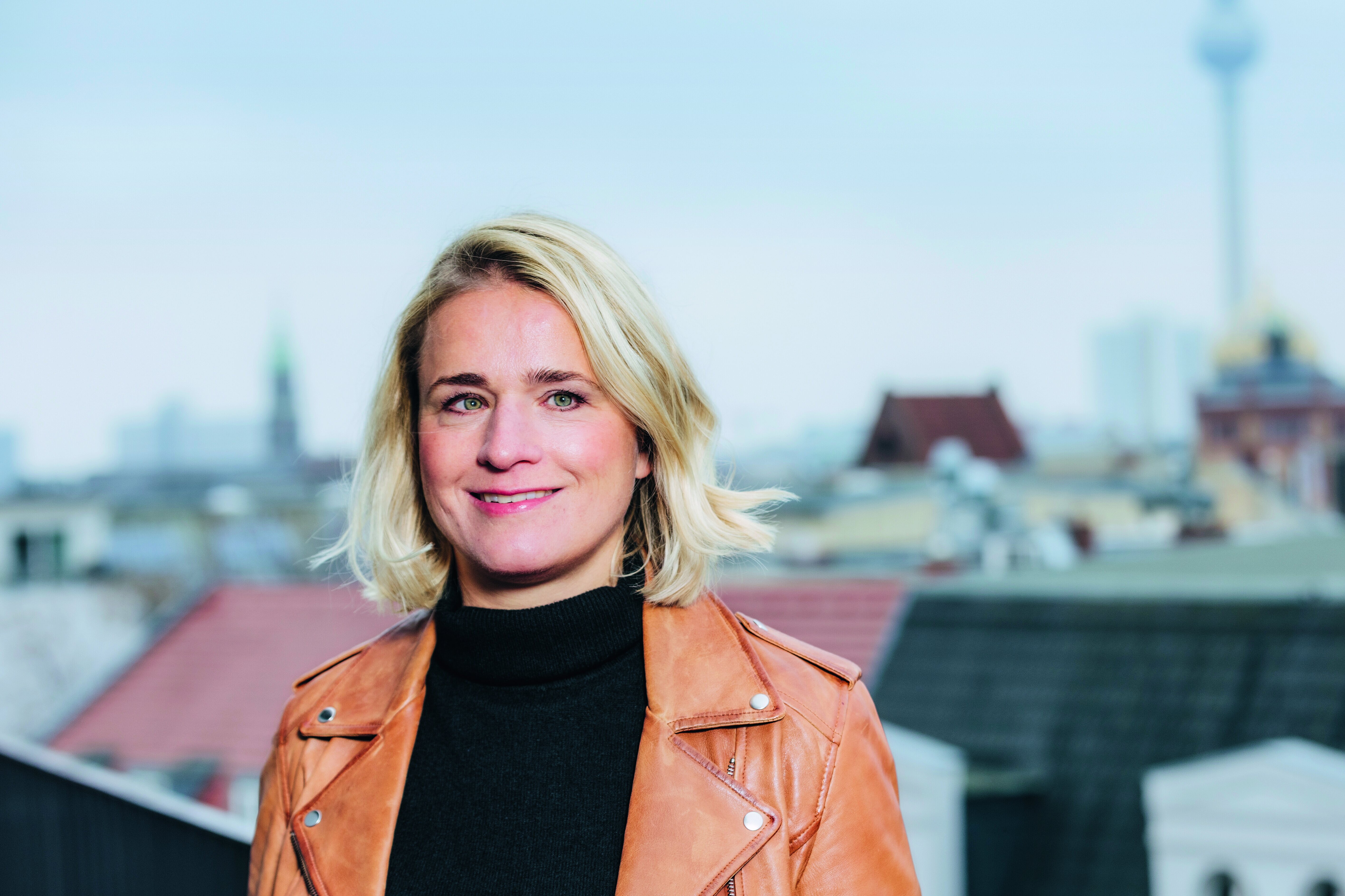 Eine Frau mit blonden Haaren und einer braunen Lederjacke steht lächelnd auf einer Dachterrasse. Im Hintergrund ist eine Stadtlandschaft mit einem Fernsehturm zu sehen.