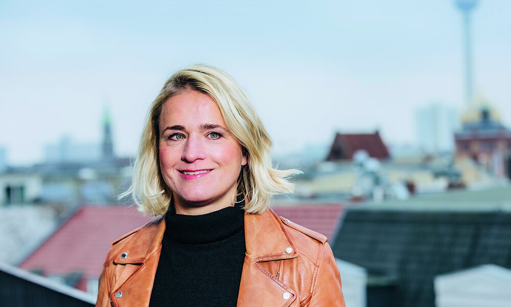 Eine Frau mit blonden Haaren und einer braunen Lederjacke steht lächelnd auf einer Dachterrasse. Im Hintergrund ist eine Stadtlandschaft mit einem Fernsehturm zu sehen.