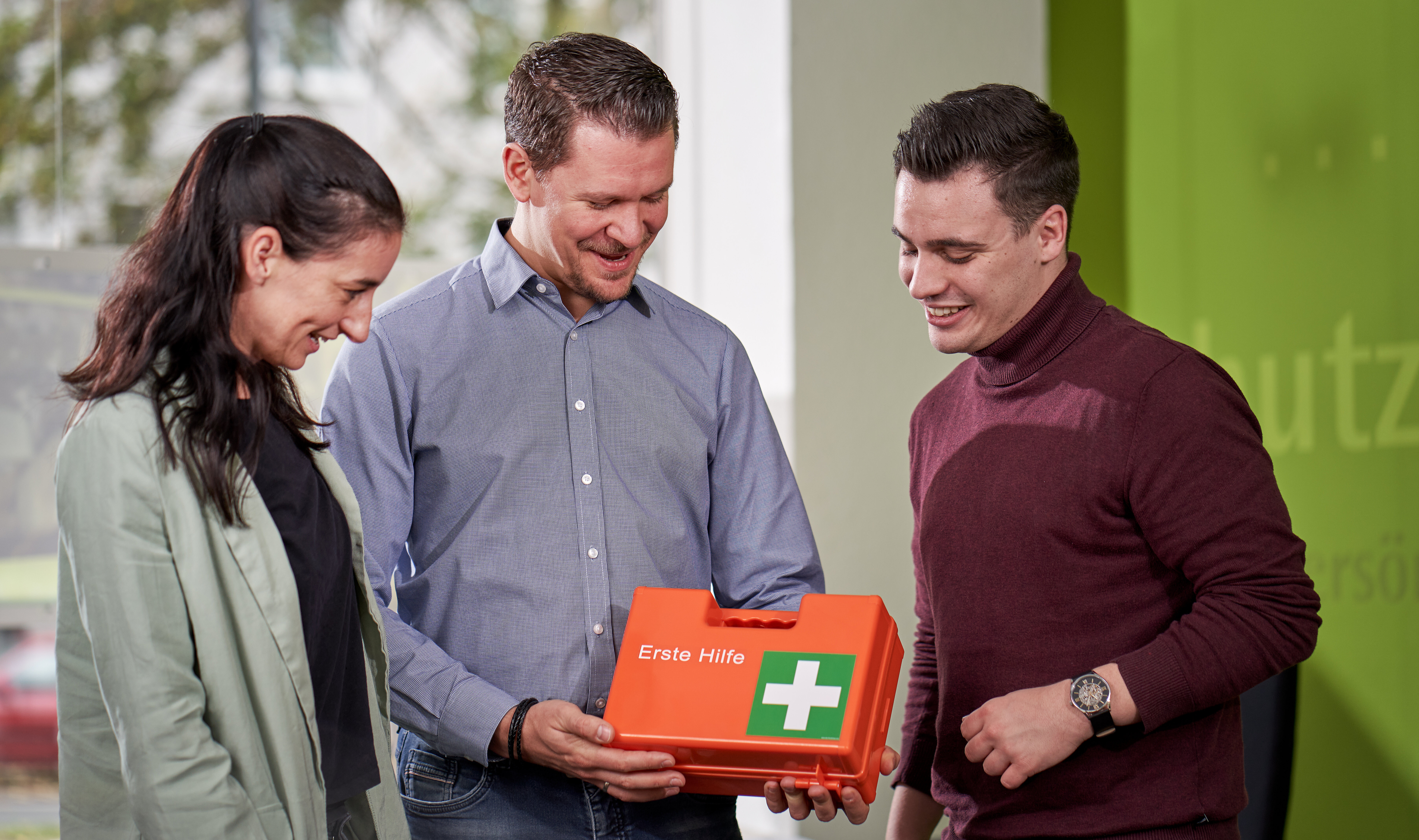 Foto: Zwei Männer und eine Frau stehen in einem Büro und schauen einen Verbandkasten an.