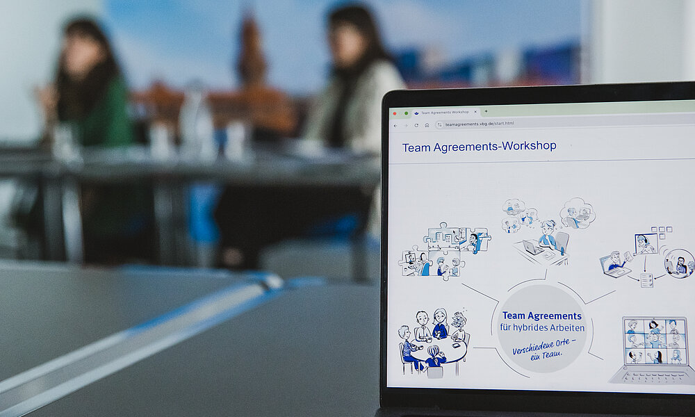Nahaufnahme des Bildschirms eines Laptops in einer Besprechung. Auf dem Bildschirm ist eine Webseite oder Präsentation mit dem Titel "Team Agreements-Workshop" und der Unterzeile "Team Agreements für hybrides Arbeiten – Verschiedene Orte – ein Team." zu sehen. Der Inhalt besteht aus Strichzeichnungen und Illustrationen, die verschiedene Aspekte von hybrider Zusammenarbeit darstellen, wie Teamtreffen vor Ort, virtuelle Meetings und Einzelarbeit. Im unscharfen Hintergrund sitzen zwei Frauen an einem Konferenztisch.
