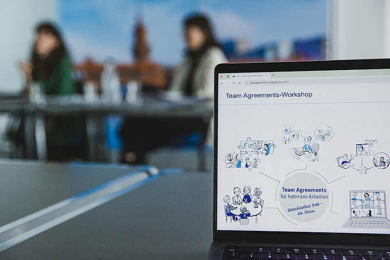 Nahaufnahme des Bildschirms eines Laptops in einer Besprechung. Auf dem Bildschirm ist eine Webseite oder Präsentation mit dem Titel "Team Agreements-Workshop" und der Unterzeile "Team Agreements für hybrides Arbeiten – Verschiedene Orte – ein Team." zu sehen. Der Inhalt besteht aus Strichzeichnungen und Illustrationen, die verschiedene Aspekte von hybrider Zusammenarbeit darstellen, wie Teamtreffen vor Ort, virtuelle Meetings und Einzelarbeit. Im unscharfen Hintergrund sitzen zwei Frauen an einem Konferenztisch.