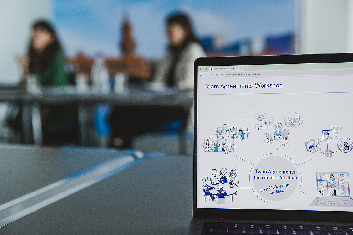 Nahaufnahme des Bildschirms eines Laptops in einer Besprechung. Auf dem Bildschirm ist eine Webseite oder Präsentation mit dem Titel "Team Agreements-Workshop" und der Unterzeile "Team Agreements für hybrides Arbeiten – Verschiedene Orte – ein Team." zu sehen. Der Inhalt besteht aus Strichzeichnungen und Illustrationen, die verschiedene Aspekte von hybrider Zusammenarbeit darstellen, wie Teamtreffen vor Ort, virtuelle Meetings und Einzelarbeit. Im unscharfen Hintergrund sitzen zwei Frauen an einem Konferenztisch.