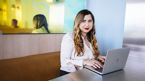 Eine Frau sitzt an einem Tisch mit einem Laptop und schaut in die Kamera. Sie trägt eine weiße Bluse und hat lange, gewellte Haare. Im Hintergrund ist ein verschwommenes Spiegelbild zu sehen.