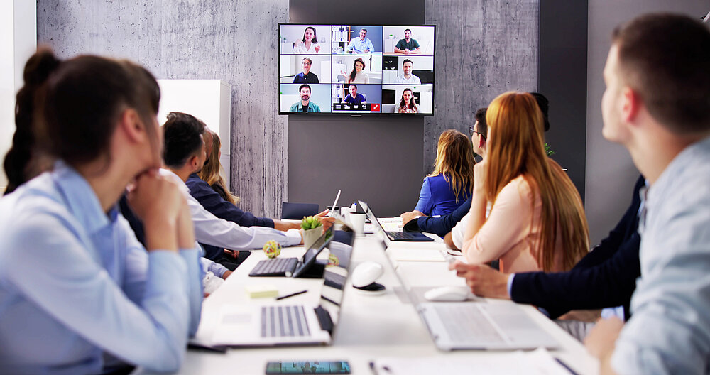 Eine Gruppe von Menschen sitzt an einem Konferenztisch und schaut auf einen Bildschirm, auf dem ein Videoanruf mit mehreren Teilnehmern zu sehen ist. Laptops und Notizen liegen auf dem Tisch.