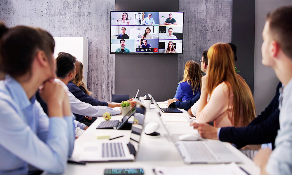 Eine Gruppe von Menschen sitzt an einem Konferenztisch und schaut auf einen Bildschirm, auf dem ein Videoanruf mit mehreren Teilnehmern zu sehen ist. Laptops und Notizen liegen auf dem Tisch.