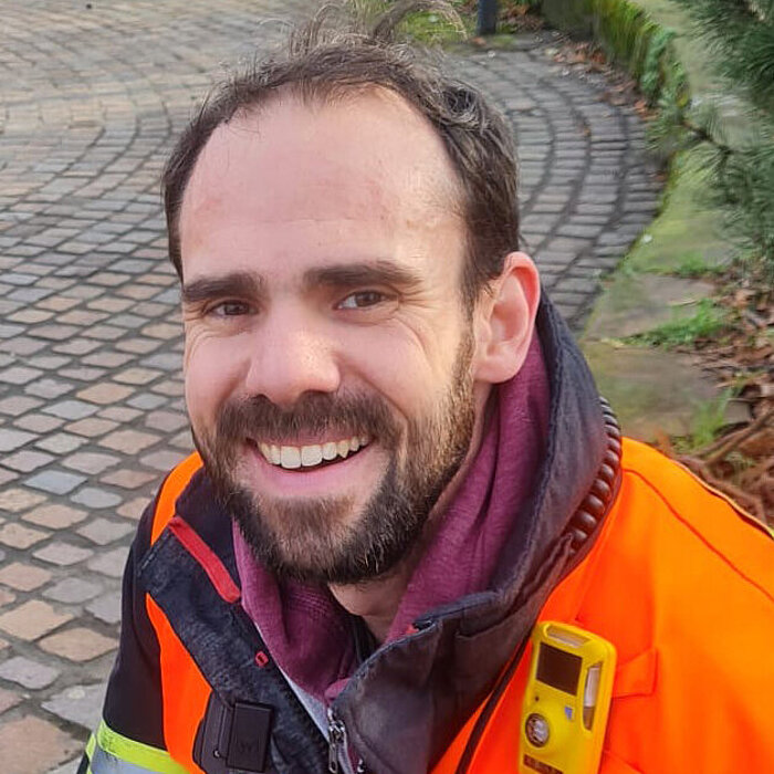 Nahaufnahme eines lächelnden Mannes mit dunklem Bart und kurzen dunklen Haaren. Er trägt eine leuchtend orangefarbene Arbeitsweste mit einem kleinen Funkgerät oder Messgerät an der Brust. Unter der Weste trägt er dunkle Kleidung. Im unscharfen Hintergrund ist ein gepflasterter Boden und etwas Grün zu sehen.