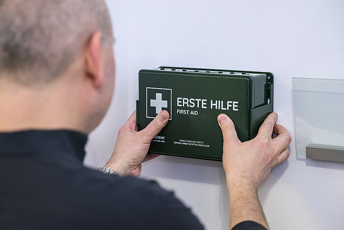 Ein kahlköpfiger Mann, der ein dunkles Oberteil trägt, befestigt eine dunkelgrüne Erste-Hilfe-Box an einer hellen Wand. Die Box trägt die Aufschrift "ERSTE HILFE" und "FIRST AID" sowie ein weißes Kreuz. Die Szene ist von hinten aufgenommen, sodass der Mann mit dem Rücken zur Kamera steht.