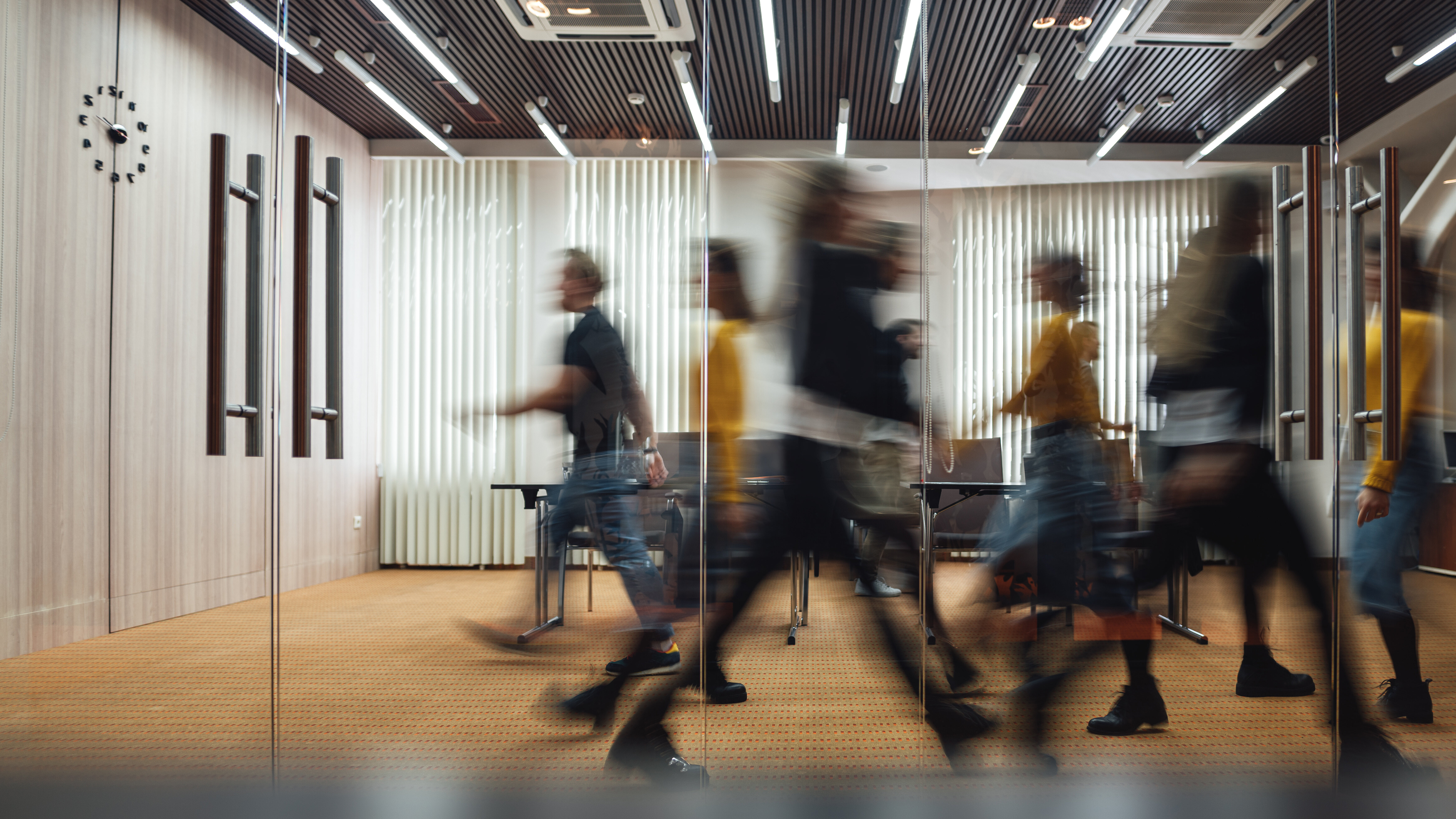 Foto: Hinter einer Glaswand in einem Büro laufen Menschen in unterschiedlichen Richtungen durch den Raum.