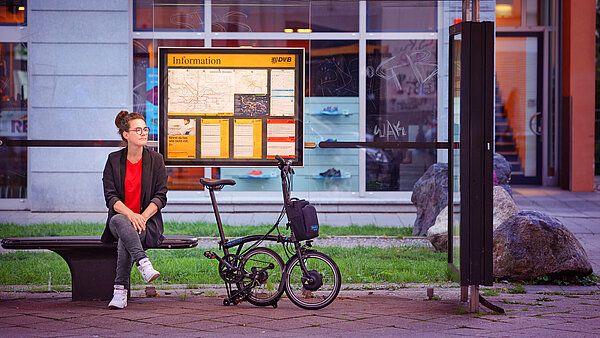 Eine junge Frau sitzt auf einer dunklen Bank im Freien vor einem modernen Gebäude. Sie trägt einen schwarzen Blazer über einem roten Oberteil, graue Hosen und weiße Turnschuhe. Sie schaut lächelnd nach links. Neben ihr steht ein zusammengeklapptes Faltrad. Hinter ihr hängt eine große, gelbe Informationstafel, wahrscheinlich mit einer Karte und Hinweisen. Der Hintergrund zeigt eine Glasfassade.